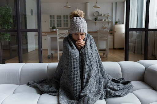 A woman is very cold inside huddling on a sofa with gray blanket and hat.